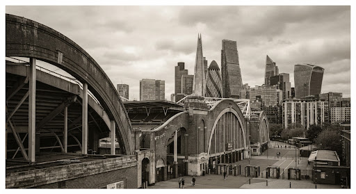 Monochrome sepia-tinged architectural view of historic European sports stadium arches meeting modern high-finance glass structures in London, minimal contrast, archival documentary fine art photography.
